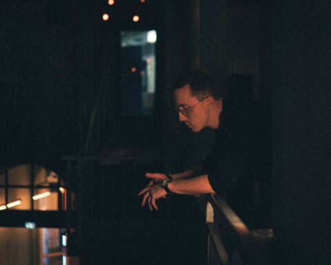 Promotional photo for "Feel It In My Bones" which sees Fred Waves standing on a balcony with his arms on the rails and he's looking down. The camera is to the side of him, so we see a darkened hallway beyond him.