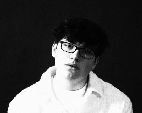 Promotional photo for "house of glass" featuring Colleen Angel, which sees a black and white head shot of Rob Eberle who is wearing a white jacket over a white t-shirt, with glasses.