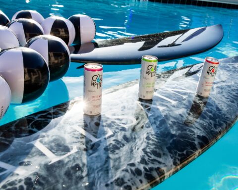 Promotional photo for AllSaints and Caliwater Sunglasses rooftop event. The photo sees two black surfboards with the AllSaints name on them, with black and white beach balls to the left, and the three Caliwater flavoured cans on the nearest board.