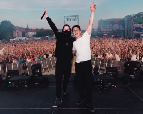Promotional photo for "Endless Summer" which sees Alan Walker and Zak Abel on stage together with their back to the festival crowd as everyone poses for the camera. Alan Walker is dressed all in black and Zak Abel is wearing a white t-shirt with black joggers.