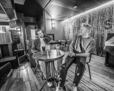 Black and white promotional photo for "How In Heaven" which sees the duo Arliston sitting at a round table on a stage in a bar, chilling in seats, drinking beer.