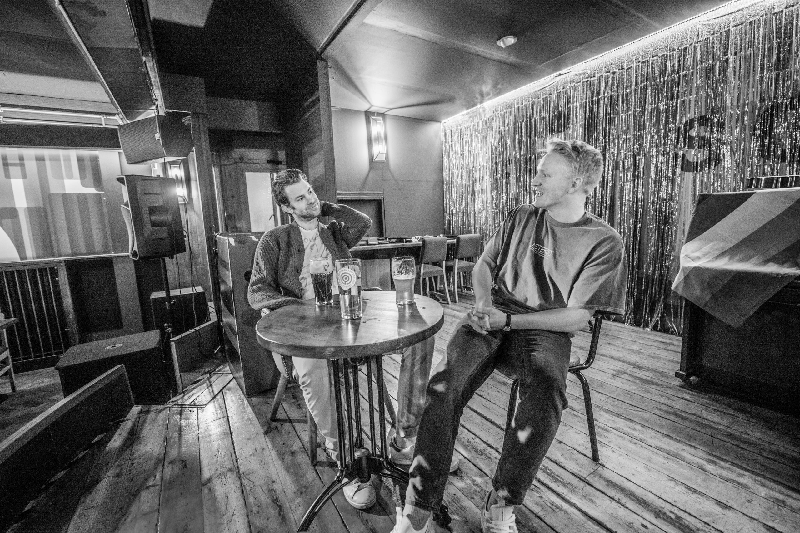 Black and white promotional photo for "How In Heaven" which sees the duo Arliston sitting at a round table on a stage in a bar, chilling in seats, drinking beer.