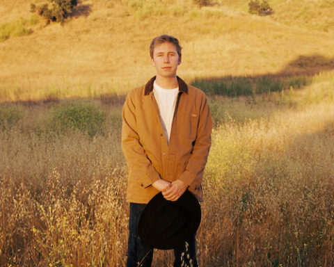Promotional photo for "Got Him!" which sees Ethan Tasch standing in a wheat field wearing a white t-shirt under a wheat-coloured jacket and blue jeans. He is holding a black cowboy hat in his hands.