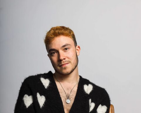 Promotional photo for "Glimpses" which sees Fox Jackson sitting on a wooden chair, wearing a black cardigan with white hearts all over it, with a low neckline, showing off his chest and silver necklace.