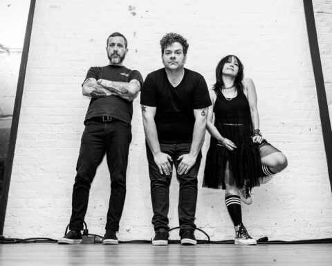 Black and white promotional photo for "Yay For Effort" which sees Greg Hoy standing between a man on the left and a woman on the right. Greg Hoy is leaning forward, wearing jeans and a t-shirt with his hands on his thighs, while the man on the left has his arms crossed over his t-shirt which he has paired with jeans, and as for the woman, she is leaning back against the wall with one foot on the wall with her knee sticking out from under her dress.