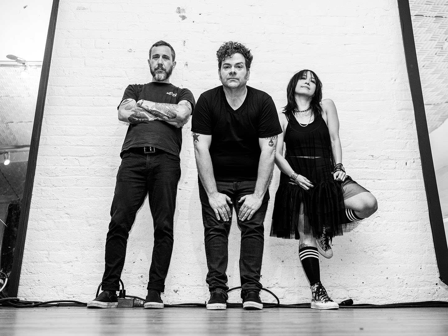 Black and white promotional photo for "Yay For Effort" which sees Greg Hoy standing between a man on the left and a woman on the right. Greg Hoy is leaning forward, wearing jeans and a t-shirt with his hands on his thighs, while the man on the left has his arms crossed over his t-shirt which he has paired with jeans, and as for the woman, she is leaning back against the wall with one foot on the wall with her knee sticking out from under her dress.