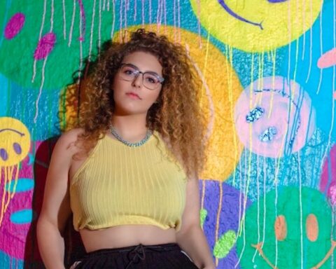 Promotional image for "Compare to Me" featuring Jamie Love, which sees Kapture leaning against a painted wall with different brightly-coloured smiley faces on it. She's wearing a yellow sleeveless top and black trousers. Her light brown afro hair reaches just past her shoulders and she completes her look with purple-rimmed glasses and a silver chain necklace.