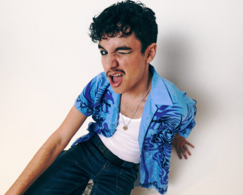 Promotional photo for "Don't Call Me At A Party" Which sees Liam Benayon wearing a white vest top under an open blue shirt with dark blue palm tree design on the sides, and some dark blue jeans. He's winking at the camera.