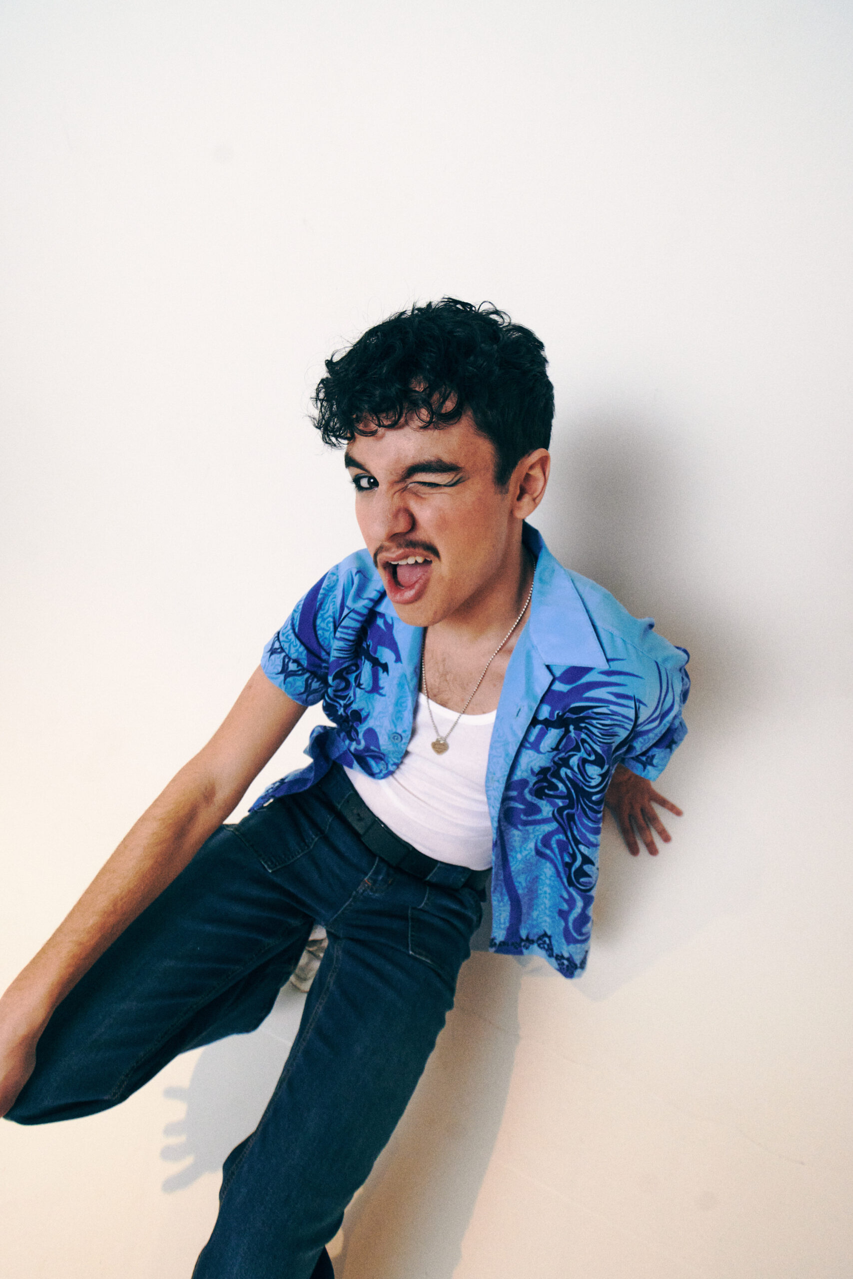 Promotional photo for "Don't Call Me At A Party" Which sees Liam Benayon wearing a white vest top under an open blue shirt with dark blue palm tree design on the sides, and some dark blue jeans. He's winking at the camera.