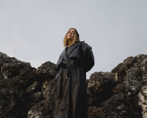 Promotional photo for "sometimes a person never comes back (but that's okay)" which sees MAVICA on a huge rock formation wearing a green trench coat and posing to the side.