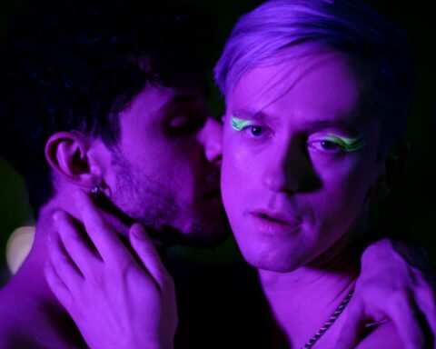 A still from the "Until Tomorrow" music video which sees Hernando Umana kissing the cheek of Magnus Riise as the singer looks at the camera with a lustful gaze. They're both shirtless, Magnus Riise's hand is on Hernando Umana's neck and the singer has luminous green eye shadow on.