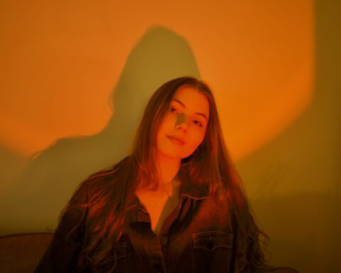 Promotional photo for "Lenox" which sees a headshot of Maya Donovan wearing a black shirt with her long brown hair falling past her shoulders. There's an orange colour filter to the image.