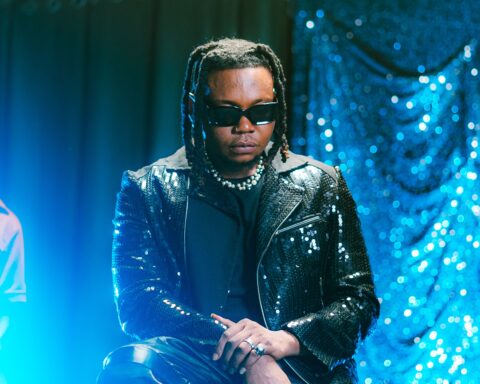 Promotional photo for "Ileke" and "Kisses" double a-side single which sees Olakira sitting in a blue-lit room wearing black leather and dark black shades.