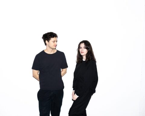 Promotional photo for "2quick2trust" which sees TANDM posing in front of a white background, with Maxine Beck-Sinderby standing to the right with Thomas Franklin standing to the left. They're both dressed in full black and slightly looking away from the camera.