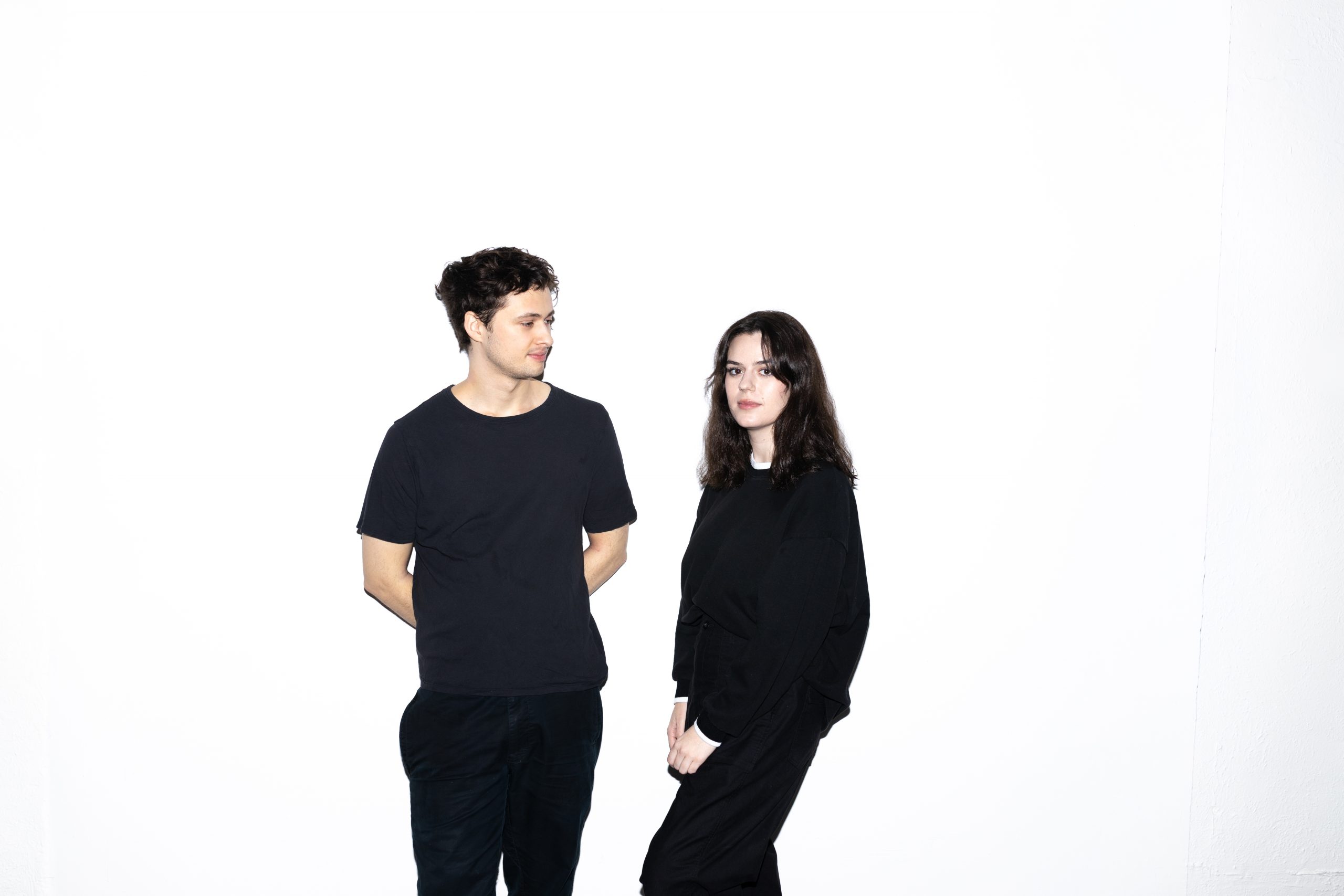 Promotional photo for "2quick2trust" which sees TANDM posing in front of a white background, with Maxine Beck-Sinderby standing to the right with Thomas Franklin standing to the left. They're both dressed in full black and slightly looking away from the camera.