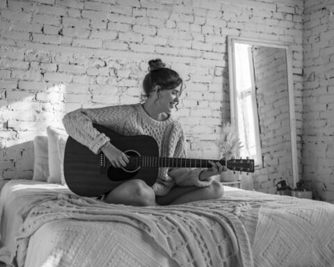 Promotional photo for "Here We Go Again" which sees a young woman, who is Victoria Staff, sitting cross-legged on a bed, playing her guitar. The photo was in black and white.
