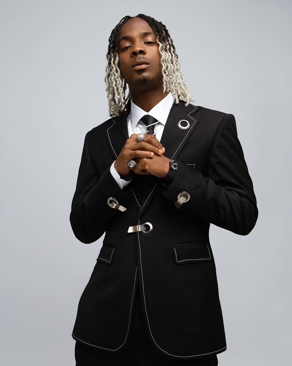 Promotional photo for "Sharpally" which sees Young Jonn posing in a photo studio, in front of a grey background, wearing a dark black suit with a white shirt and a black tie, with his dreadlock hair two-tone with blonde half-way while the rest is black to his scalp. His hair is shoulder length and parted down the middle. He is holding his hands together out in front of his chest.