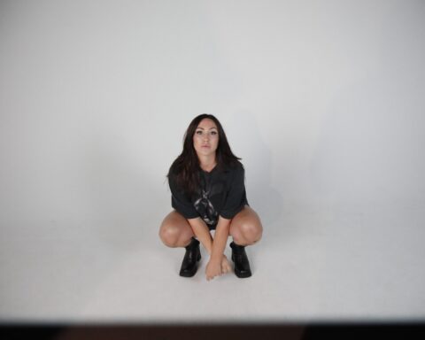 Promotional photo for "Die For Love" which sees Alyssa Reid in a white room, crouching down to the floor with her legs bare and wearing a black band t-shirt and black combat boots.