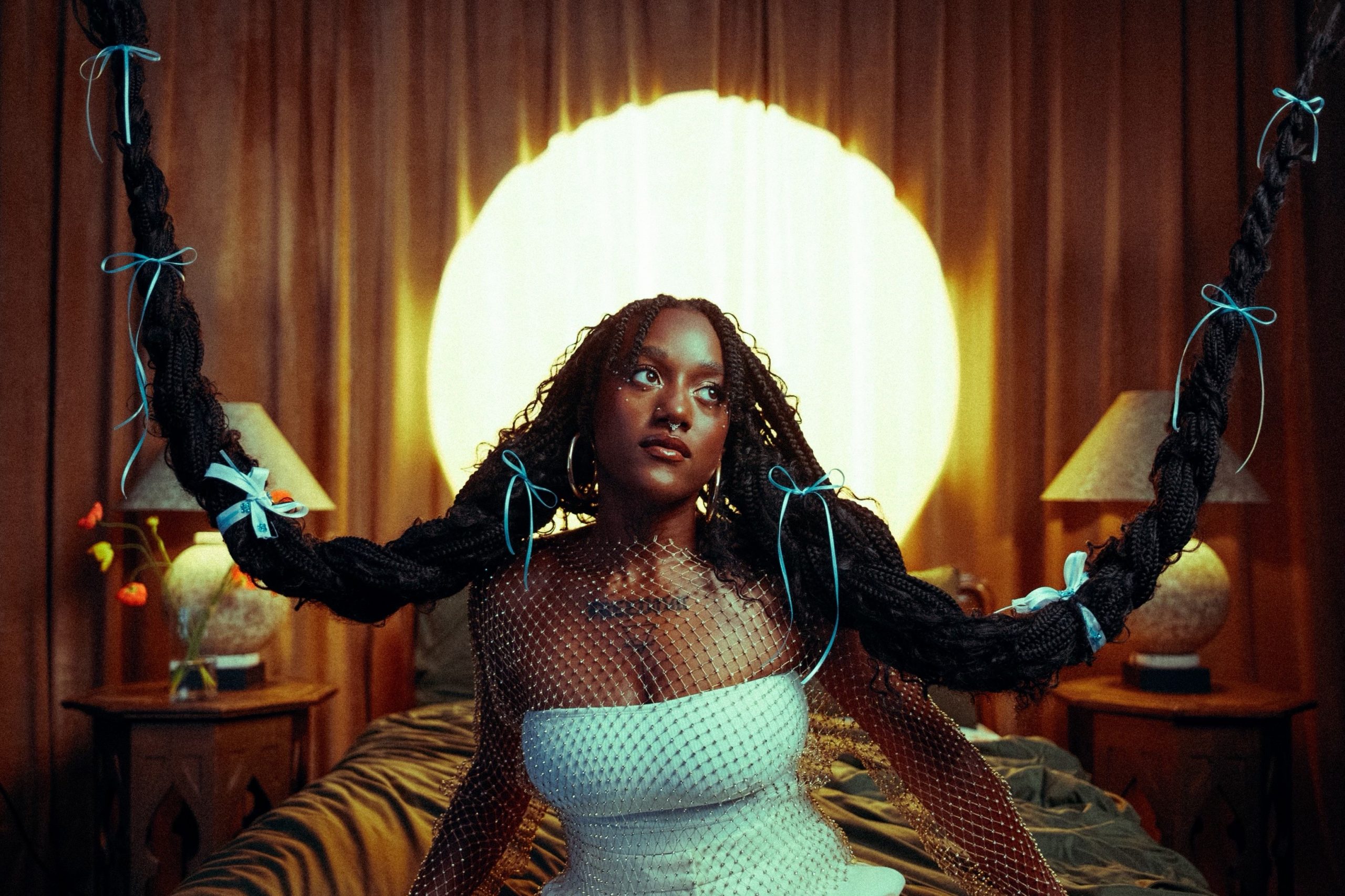 Promotional photo for "Mirror" which sees Aryeè The Gem sitting on a bed wearing a white dress and a diamante netting top, with her long black hair in two bunches which are braided and are attached to the roof by their ends, to create two plait swings, reminding us of a black-haired Rapunzel.