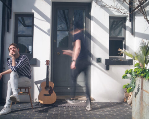 Promotional image for "C'est La Vie" which sees BANNERS sitting on the left side, in front of a house as a blurred man walks through the front door.