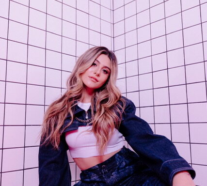 Promotional photo for "ptxd" which sees Casey Baer with blonde hair and slightly brown roots that flow past her chest, posing at the camera in a corner of a room with square patterned walls. She is wearing a fashionable denim halter top over a white crop top, paired with matching-denim trousers.
