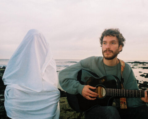 Promotional photo for "Gently, Dreamer" which sees Cayo Coco sitting down with a guitar while wearing a sea-green sweater, with the sea behind him. To his right is a person with a sheet over them like a ghost.