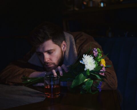 Promotional photo for "When He Was Me" which sees Charlie Rogers sitting at a table with his head on his arms which are resting on the table He has flowers in his hands and has a sad face.
