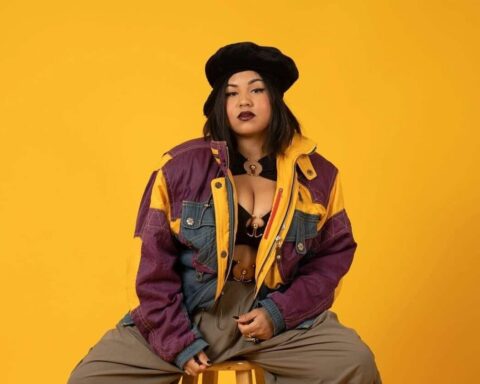 Promotional photo for "House Party" which sees Class Vee sitting on a stool in front of a bright yellow backdrop, wearing grey cargo trousers and a purple patched coat with less squares on it, with a black bra. She is also wearing a black French beret hat.