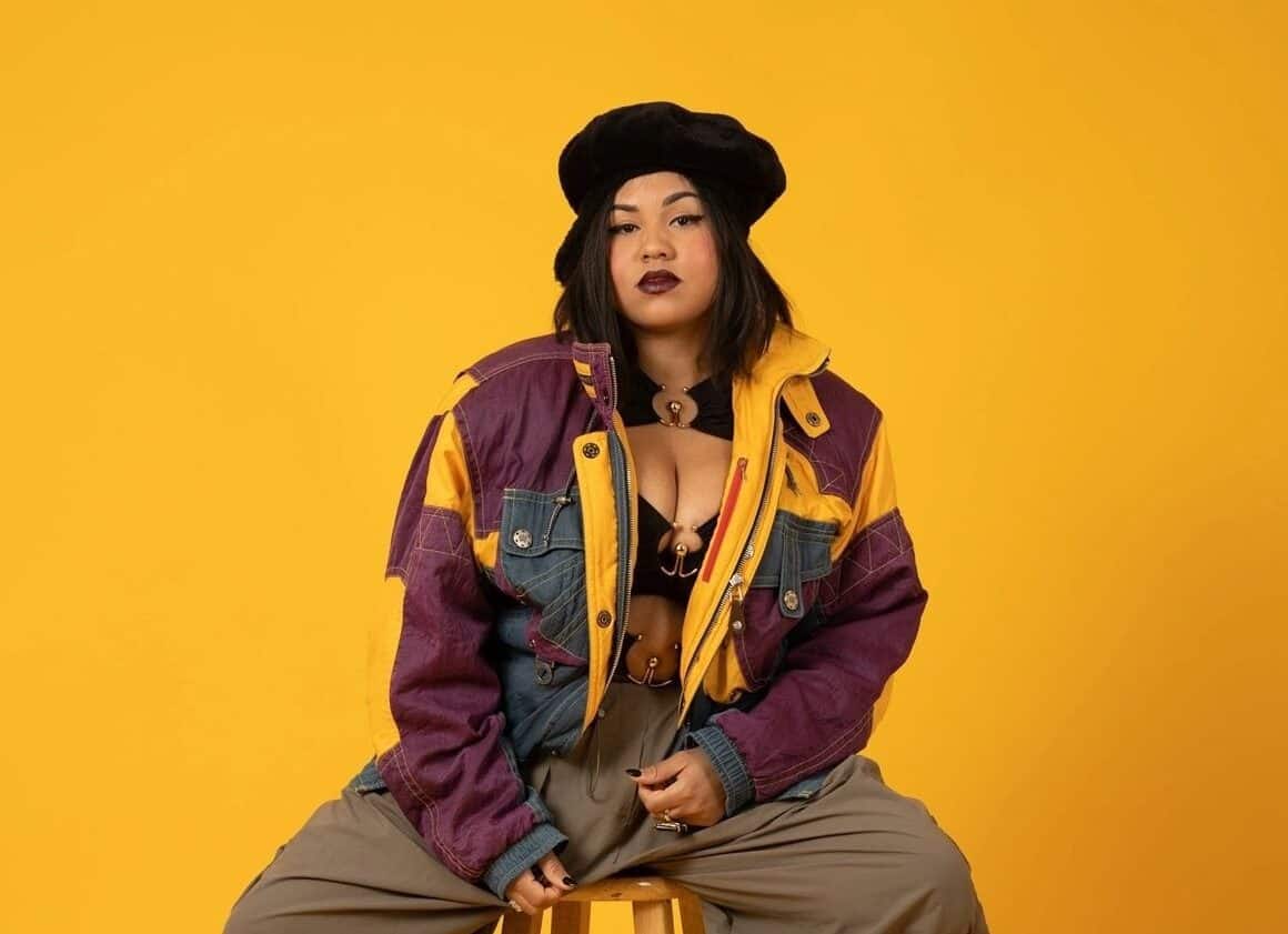 Promotional photo for "House Party" which sees Class Vee sitting on a stool in front of a bright yellow backdrop, wearing grey cargo trousers and a purple patched coat with less squares on it, with a black bra. She is also wearing a black French beret hat.