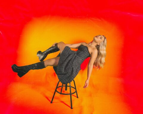 Promotional image for the music video to "Growing Pains" that sees Connie Talbot lying on a stool wearing a black dress with black knee-high boots. Her golden-blonde hair is falling down to the ground and she's balancing on the stool with her back. The background is bright orange just like the music video.