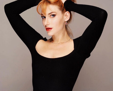 Promotional photo for "Gasoline" that sees Coyote Eyes posing for a photo shoot in front of a grey background, wearing a black square-collared long-sleeved top. She's holding her auburn hair up with both her hands.