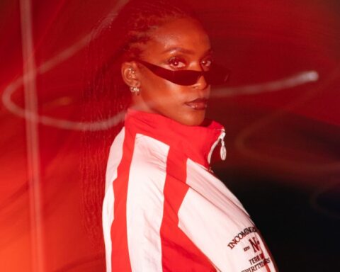 Promotional photo for "Mo Farah" which sees DEJA wearing a red and white jacket zipped to the top, with thin dark shades on. She's standing to the right with her head turned towards the camera and her long braided hair flying behind her.