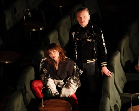Promotional photo for "Us Against The World" EP which sees the Little Lies duo in a theatre stalls posing for a photograph. Anna Maria Espinosa is sitting down, sidewards in one of the green chairs, wearing black, while Mikael Nordgren is standing behind her, also dressed in black.