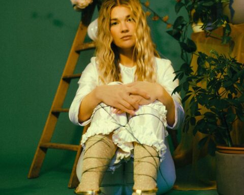 Promotional photo for "Where I Go" which sees PEARL with her knees to her chest and her arms around her legs, wearing a white shirt and matching trousers with gold boots that match her wavy blonde hair. She's sitting in a royal green-painted room.