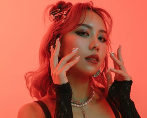 Promotional photo for "Play With You" which sees a headshot of JVNA with her hands on framing her face while her red shoulder-length hair is gathered behind her. She's lit in an amber glow.