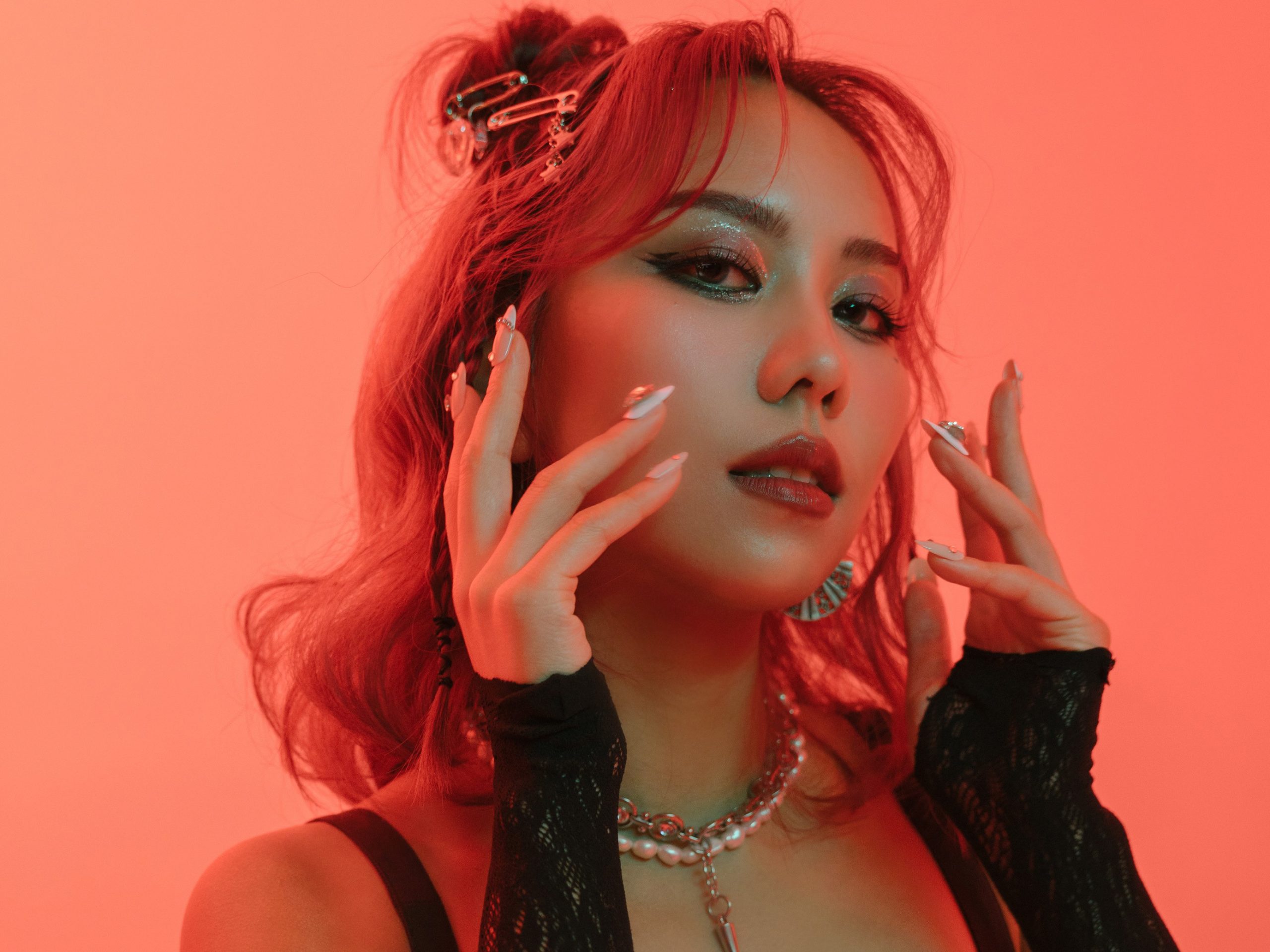 Promotional photo for "Play With You" which sees a headshot of JVNA with her hands on framing her face while her red shoulder-length hair is gathered behind her. She's lit in an amber glow.
