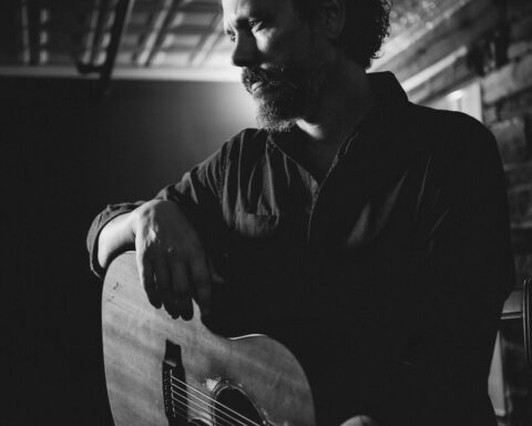Promotional photo for "Big Love" with Wildo DM, which is a black and white photo of John Paul White looking to the left, sitting down with his guitar sat on his lap and one arm holding the body of the guitar.