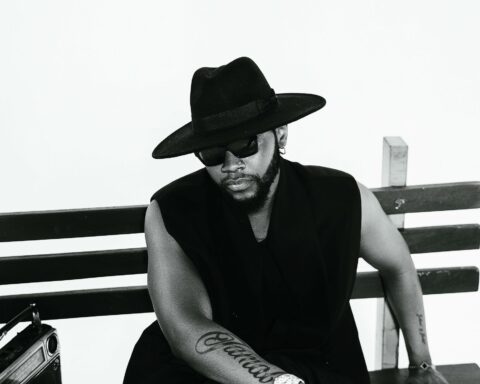 A black-and-white promotional photo for "Maverick" which sees Kizz Daniels sitting on a bench wearing a wearing a gilet jumper, with a broad-brimmed hat and dark shades, with one arm across his legs showing off his tattoo that says "Jamal" in cursive text all along his forearm.