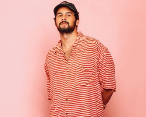 Promotional photo for "Sky Father" in collaboration with Antdot and Beacon Bloom, which sees Maz (BR) standing in front of a baby pink backboard, wearing a salmon-white checkered shirt and a flat cap.