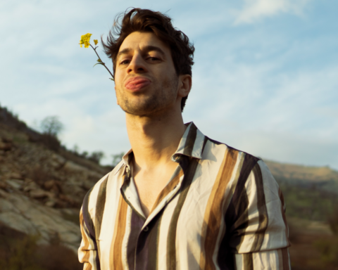 Promotional photo for "Unconditional" which sees Mike Ruby sticking his tongue out, with a daisy in his hair, and he is wearing a vertical-striped white and brown shirt. The countryside hills can be seen in the background.