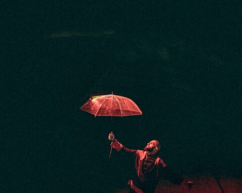 Promotional image for "Reckless" which sees a long shot of Moh Flow lit in a red spotlight, holding out an umbrella.