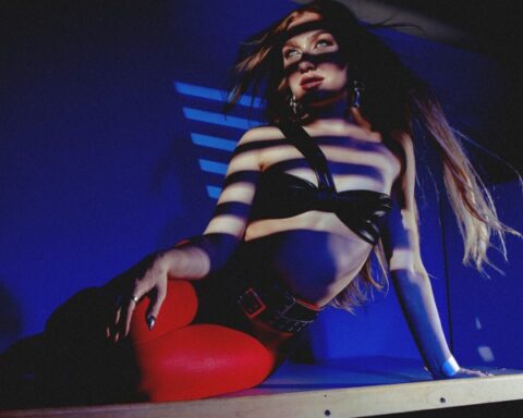 The single cover artwork for "HEARTBREAK ON REPEAT" which sees Olivia Lunny wearing red leather pants and a black bra as she lies on a metal counter with a wind machine flowing her hair behind her, in front of a blue background.