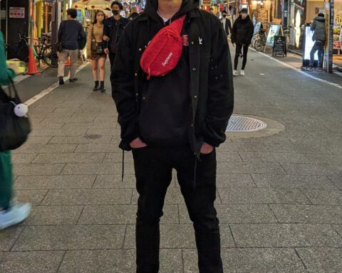 Promotional photo for "Ocean Drive" which sees Poet Initiative standing on a pedestrian shopping street with the shop lights illuminating the street. He is wearing all black, has his hands in the pockets of his black skinny jeans, and he has a red cross-body bag over his black hoodie.